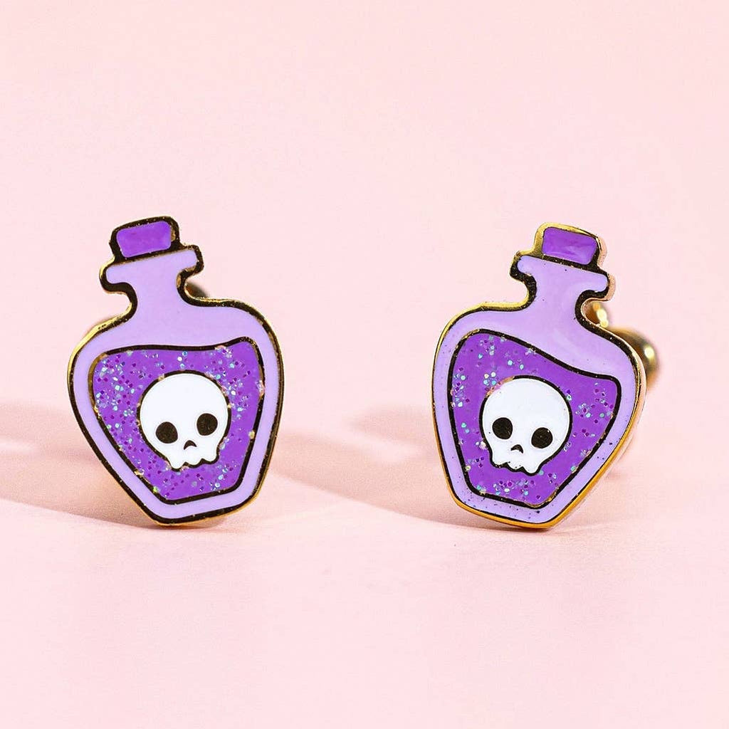 A pair of skull potion bottle stud earrings in purple and lavender with glittery purple liquid details, gold-tone metal outlines, and white skulls on a light pink background.