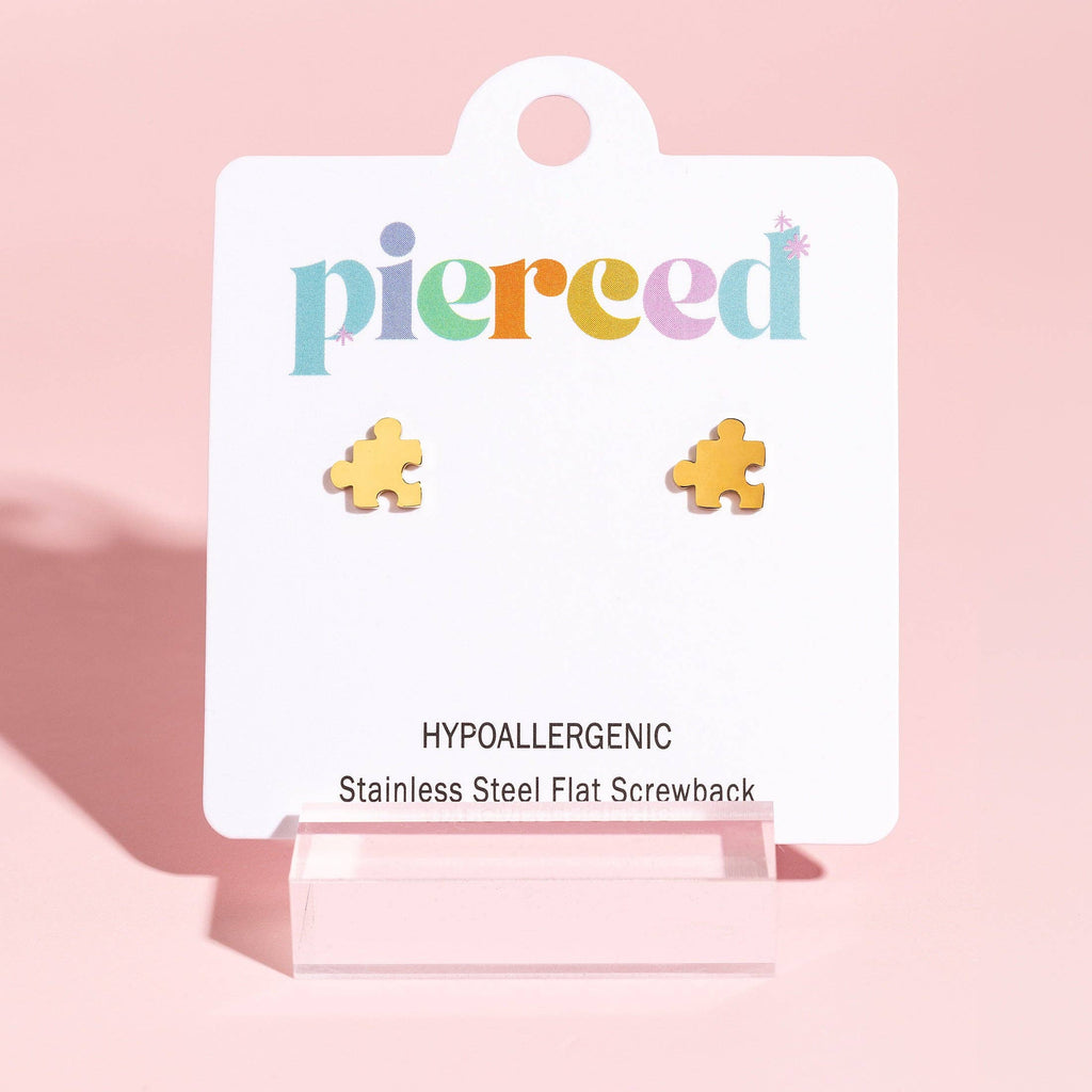 A pair of small gold puzzle-piece stud earrings displayed on a white branded card that reads 'pierced' and 'Hypoallergenic Stainless Steel Flat Screwback,' set against a soft pink background.