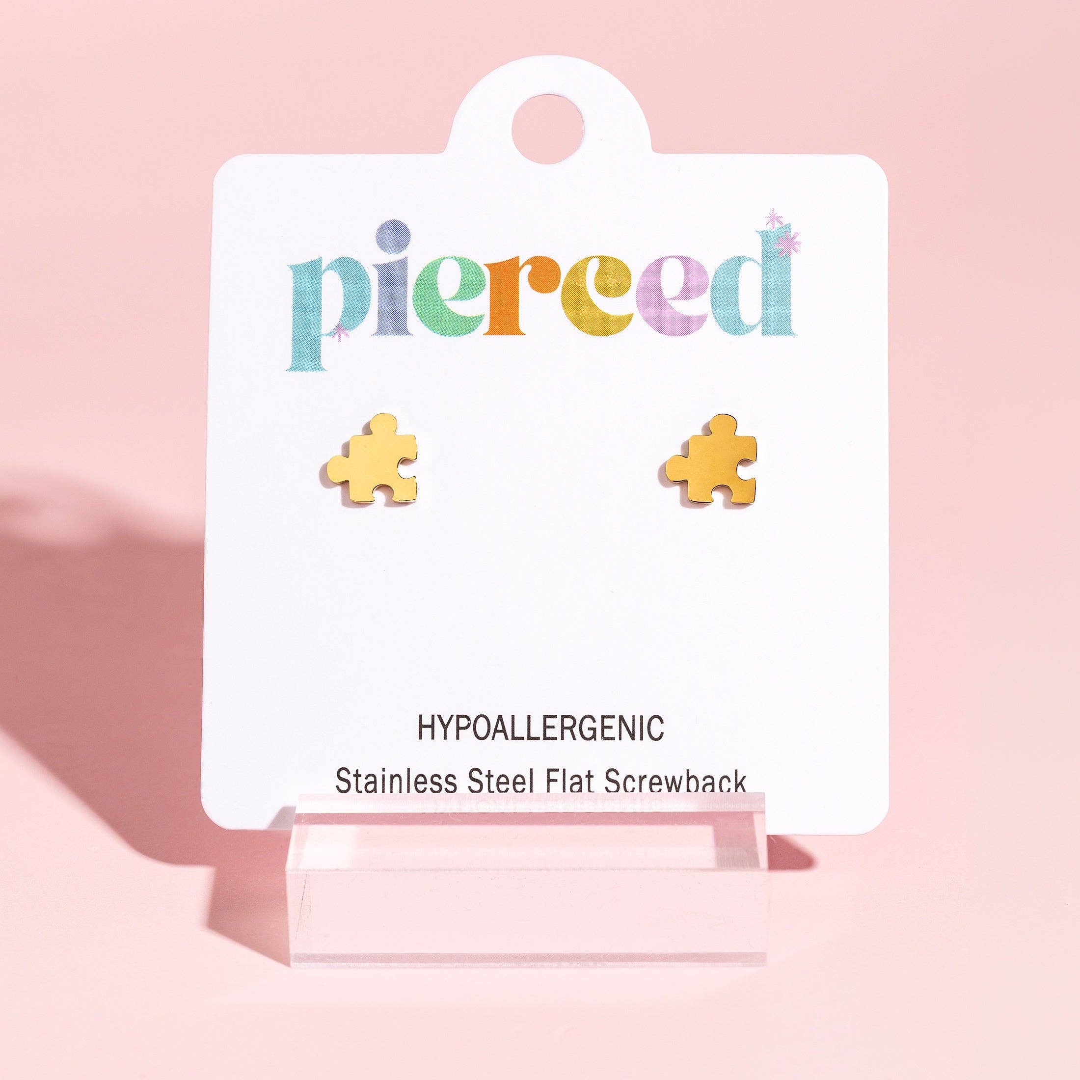 A pair of small gold puzzle-piece stud earrings displayed on a white branded card that reads 'pierced' and 'Hypoallergenic Stainless Steel Flat Screwback,' set against a soft pink background.