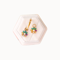 A pair of gold-tone flower-shaped earrings with multicolor enamel petals (green, blue, purple, orange) and white centers, displayed on a white hexagonal velvet jewelry box against a light off-white background.