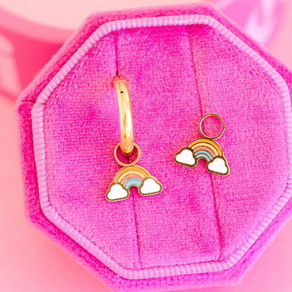 Gold hoop earrings with rainbow and cloud charms on a pink hexagonal pillow.