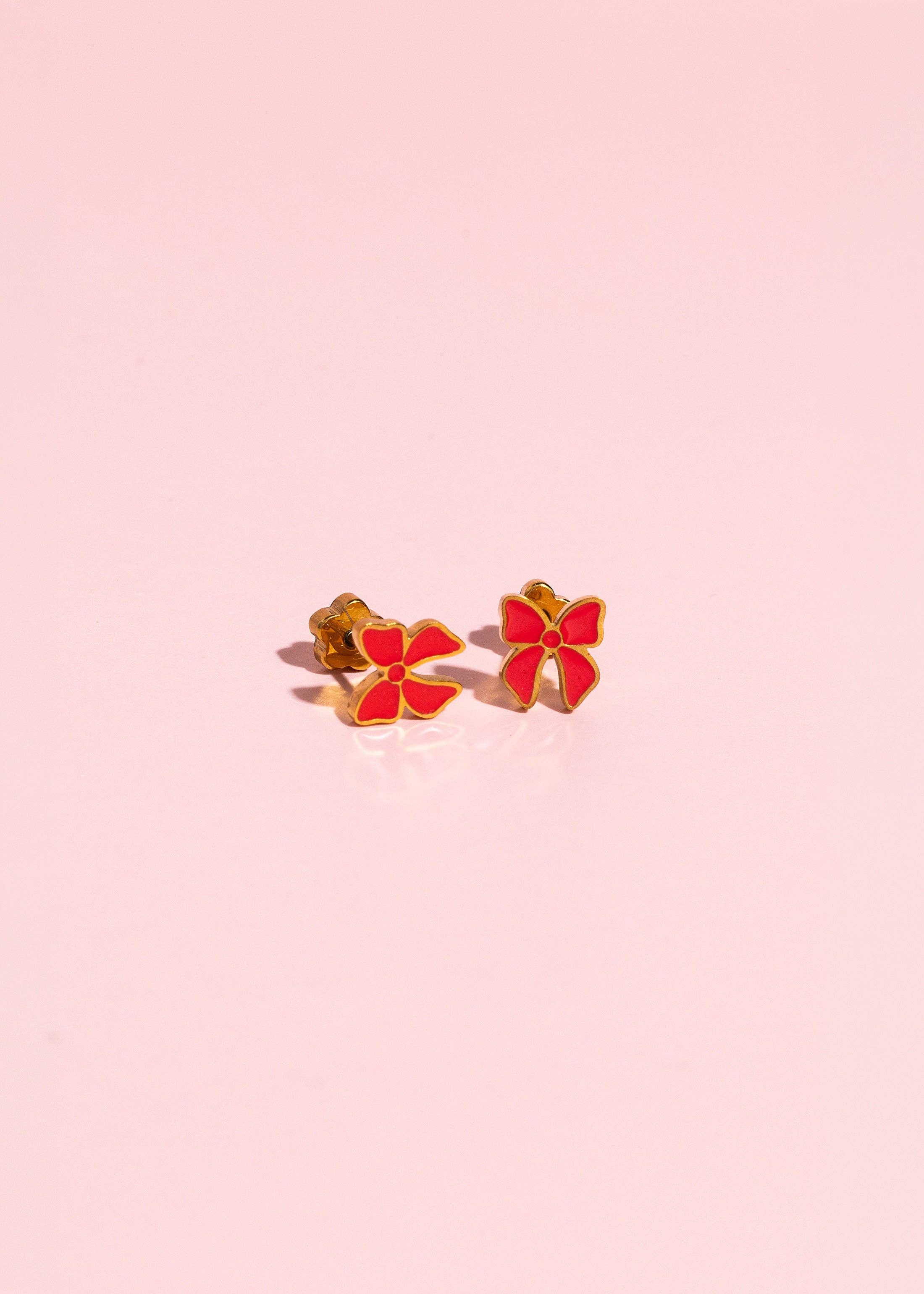 A pair of small gold-tone stud earrings shaped like red four-petal flowers, displayed on a soft pink reflective surface.