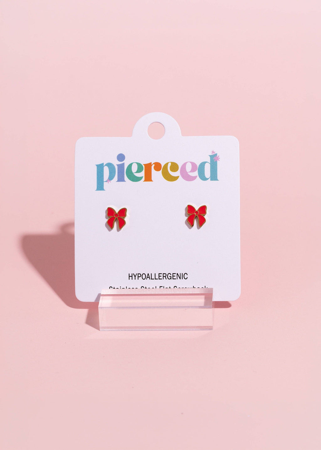 A pair of small red bow stud earrings displayed on a white card labeled 'pierced' against a soft pink background.
