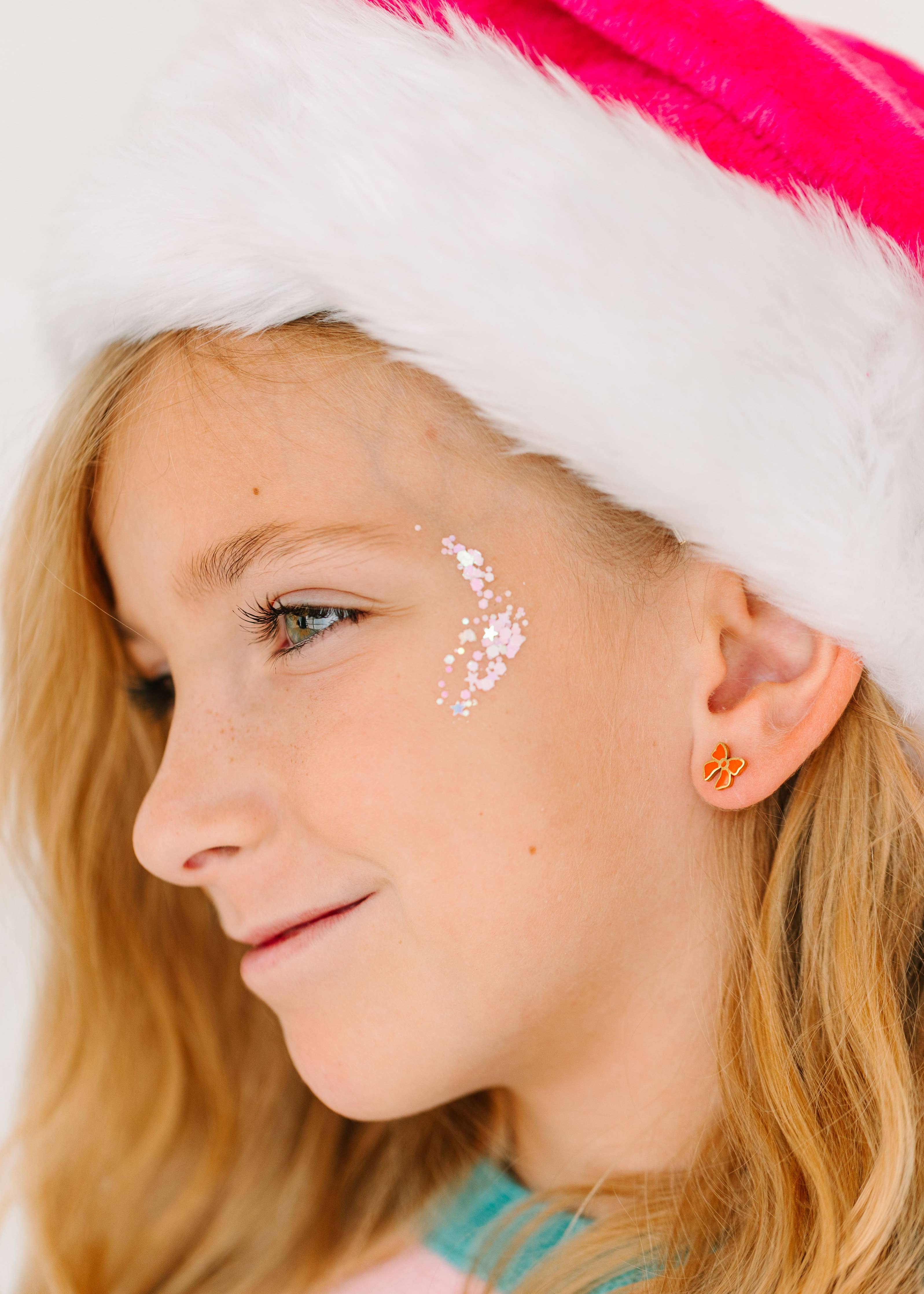 A close-up side profile of a young girl wearing a white Santa hat with a bright pink band, white faux-fur trim, and a small orange flower-shaped stud earring.