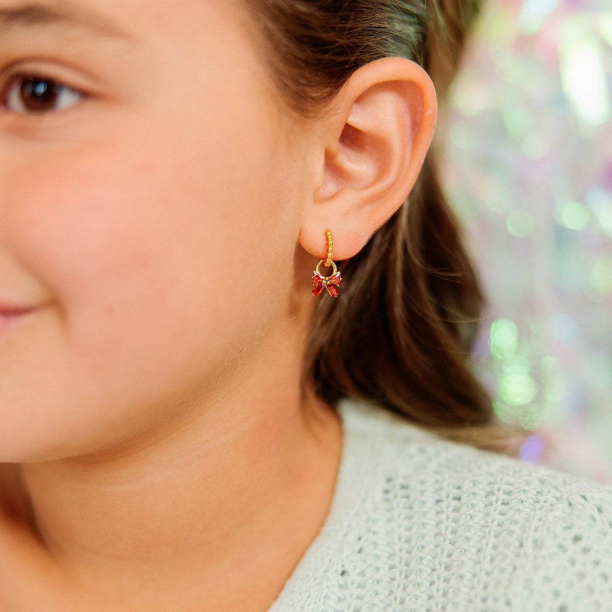 A close-up of a child's ear wearing a small gold hoop earring with dangling red bow charms, against a softly blurred background.