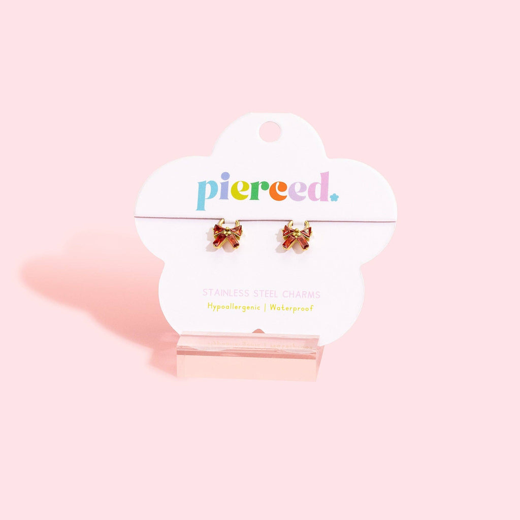 A pair of small bow-shaped stainless steel charms with red and yellow accents, displayed on a white cloud-shaped Pierced brand card stand against a solid light pink background.