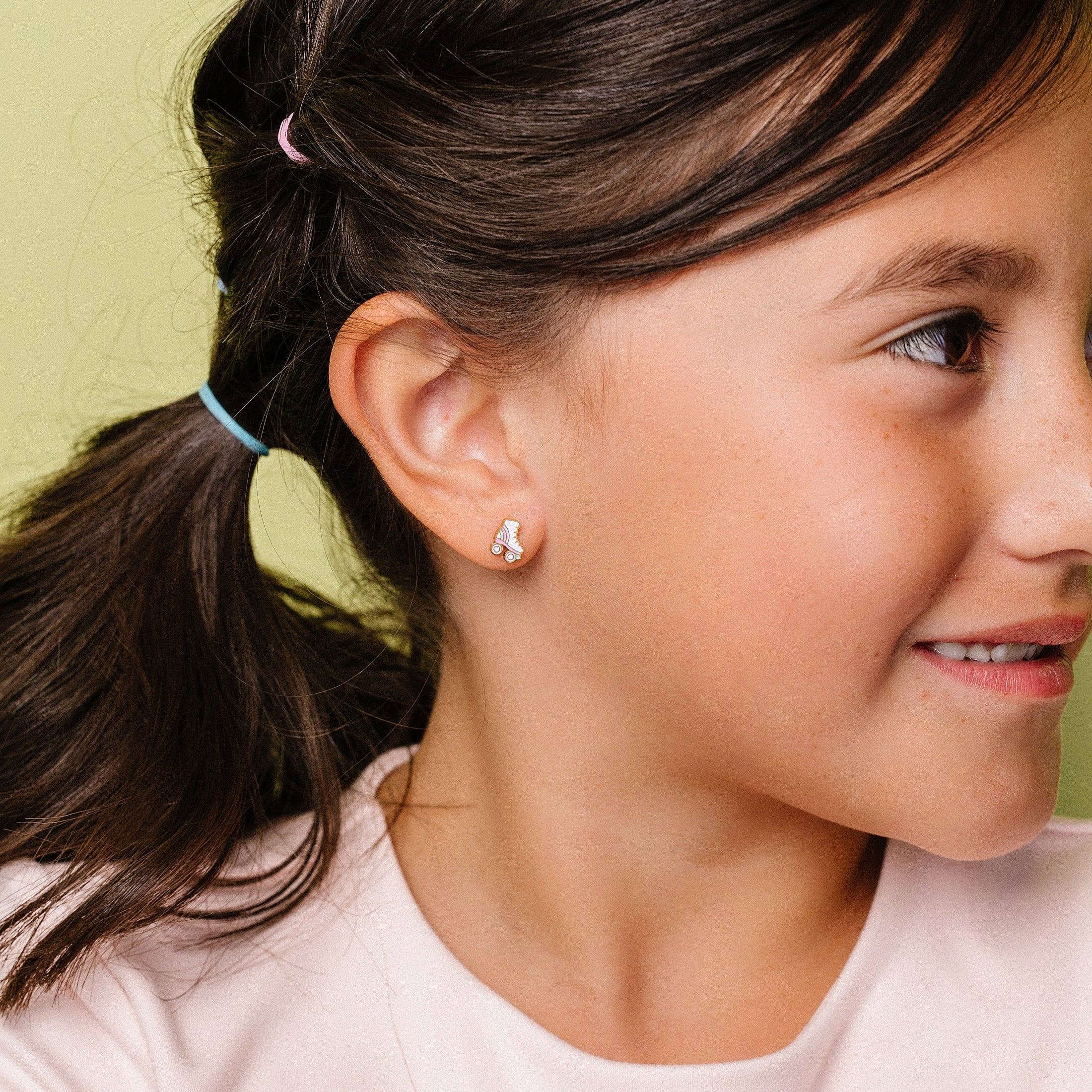 A close-up of a young girl with dark brown hair in a ponytail, wearing a small silver crescent moon stud earring, against a pale yellow-green background.