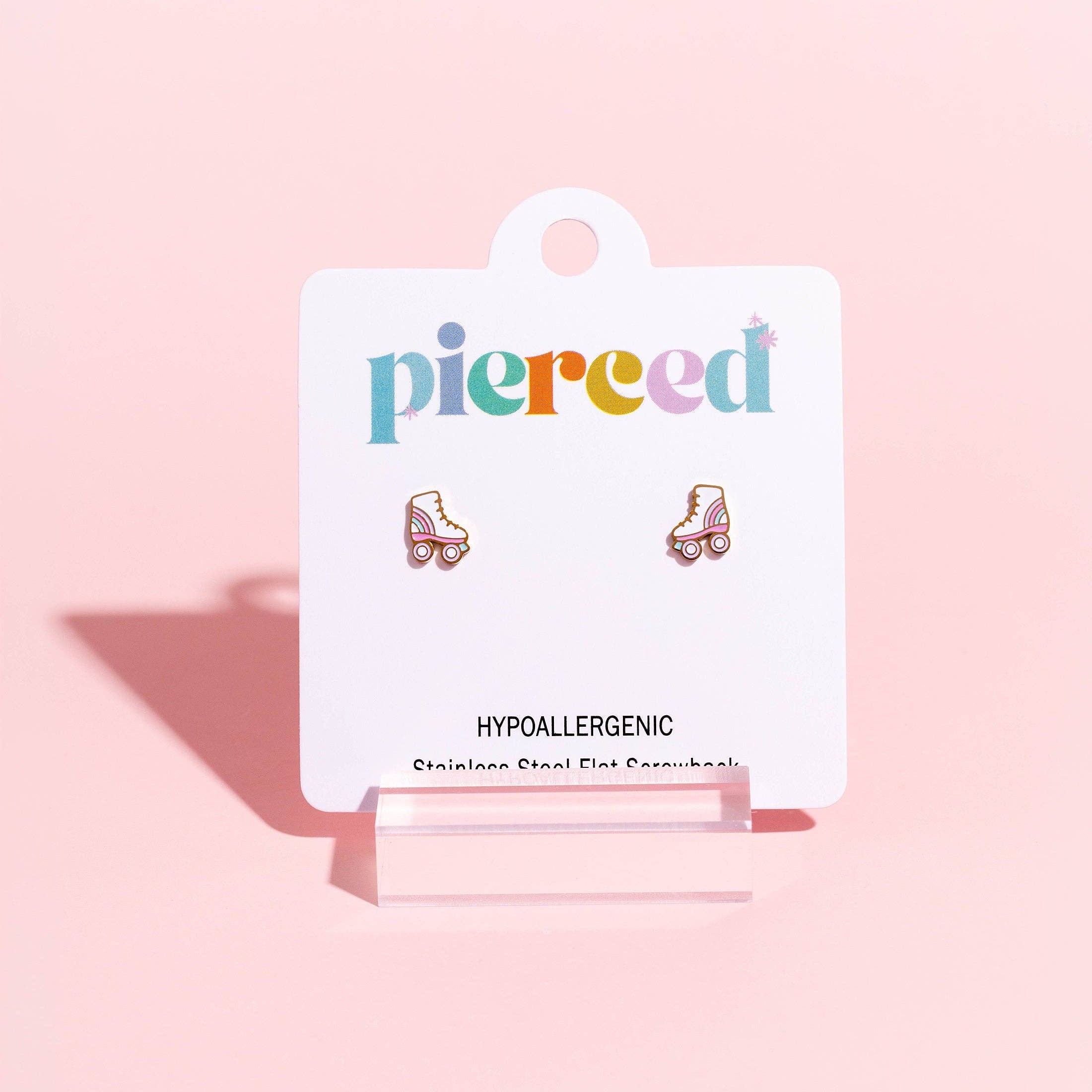 A pair of small roller skate stud earrings displayed on a white card backing that reads 'pierced' and 'HYPALLERGGENIC,' set against a soft pink background.