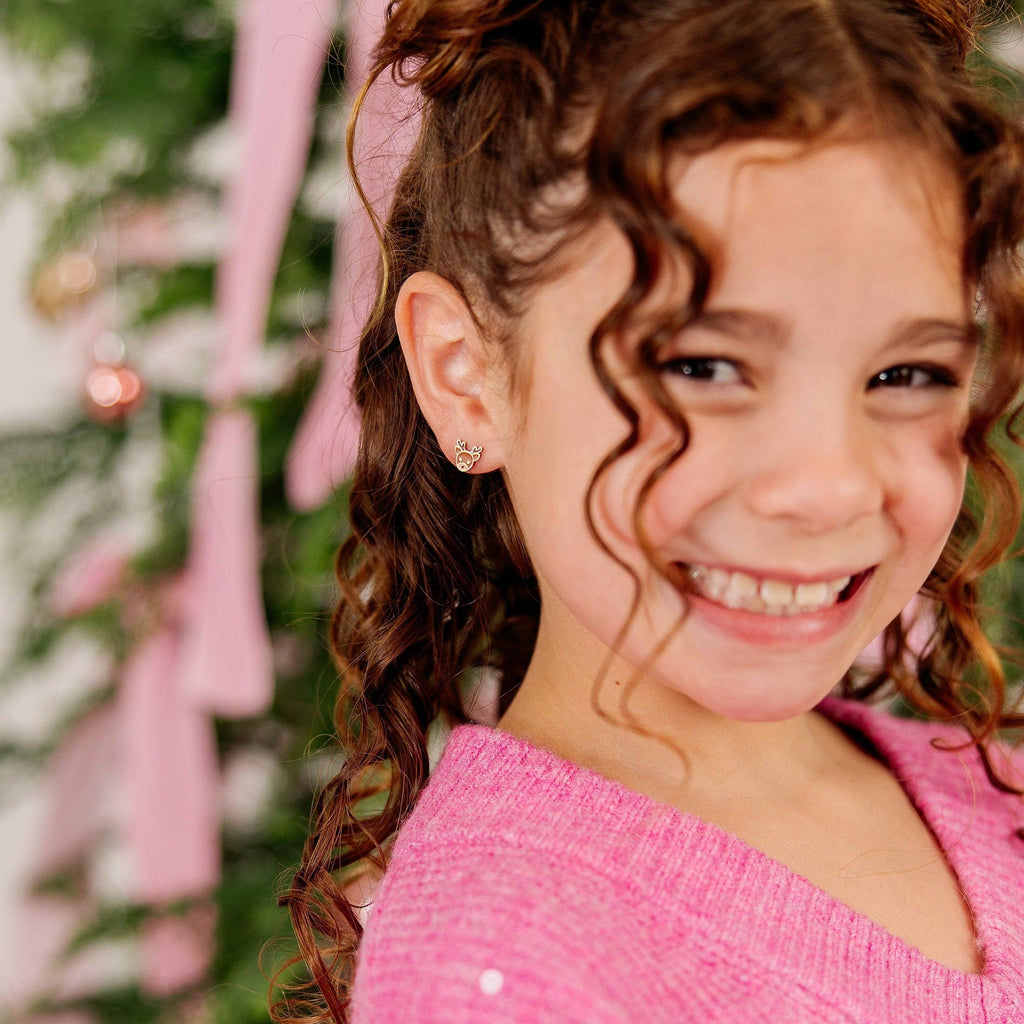 A young girl with curly brown hair styled in two buns smiles at the camera, wearing a pink knit sweater with sequins. The background is softly blurred with pink ribbons and greenery.