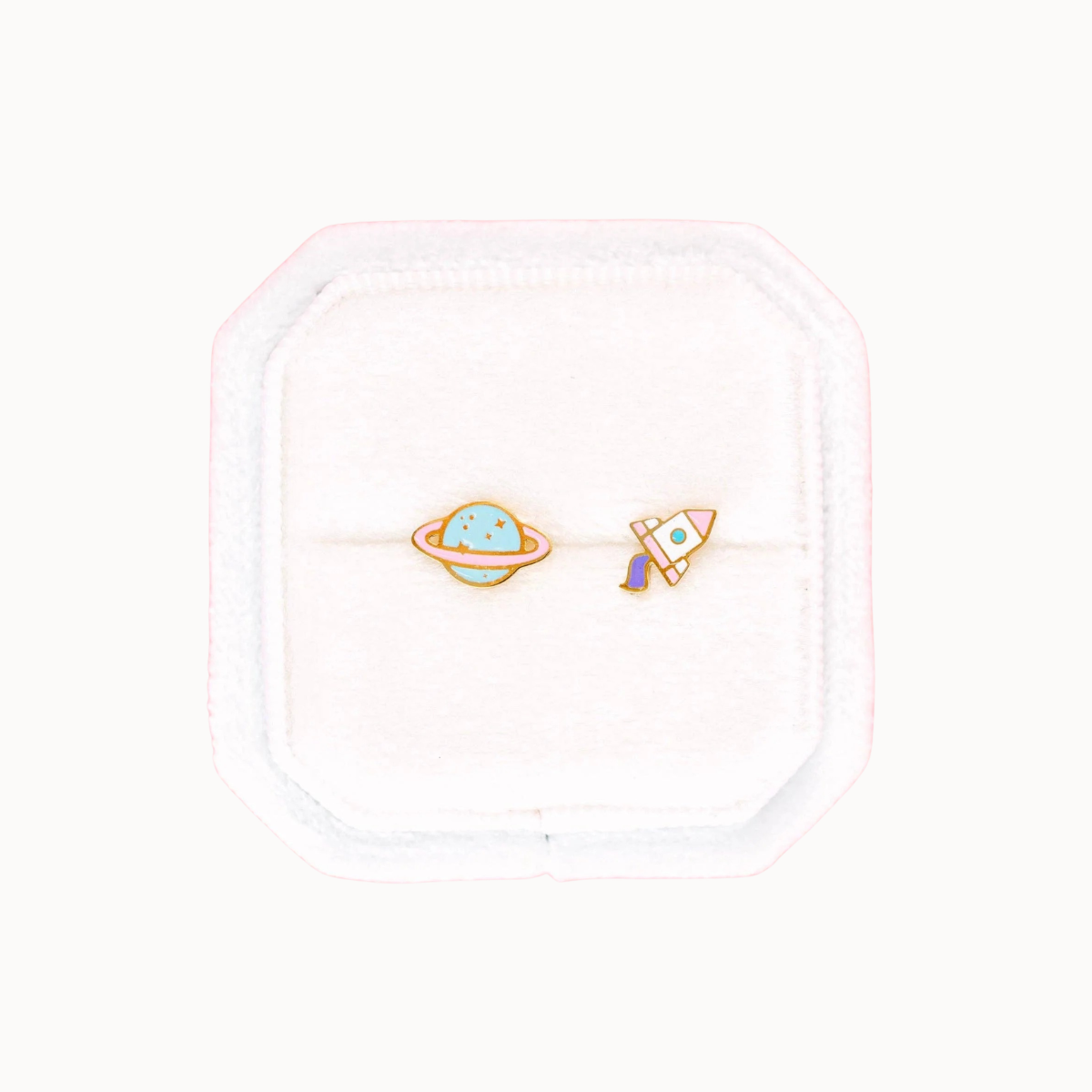 A pair of small space-themed stud earrings displayed on a white velvet jewelry box. One earring features a light blue planet with a ring, and the other features a small rocket ship.