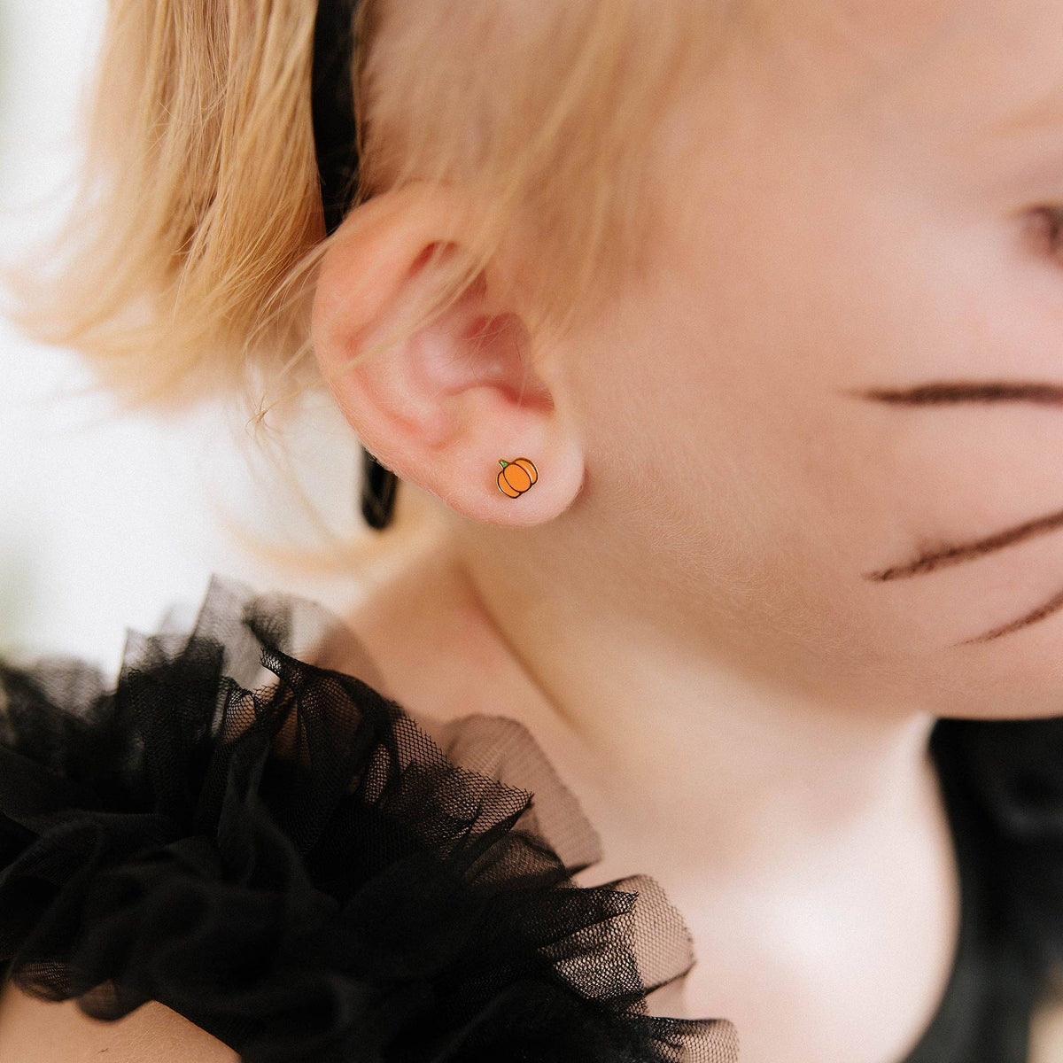 A close-up of a child's ear wearing a small round orange stud earring, with blonde hair, a black headband, and a black ruffled costume top visible.