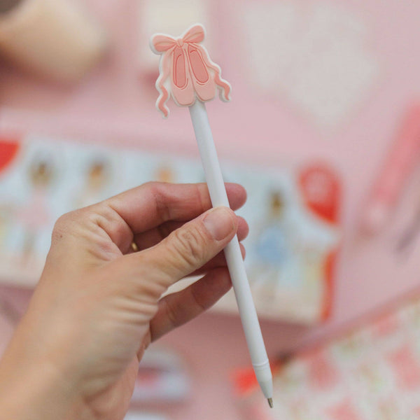 Hand holding a white pen with a pink ballerina clip against a blurred background