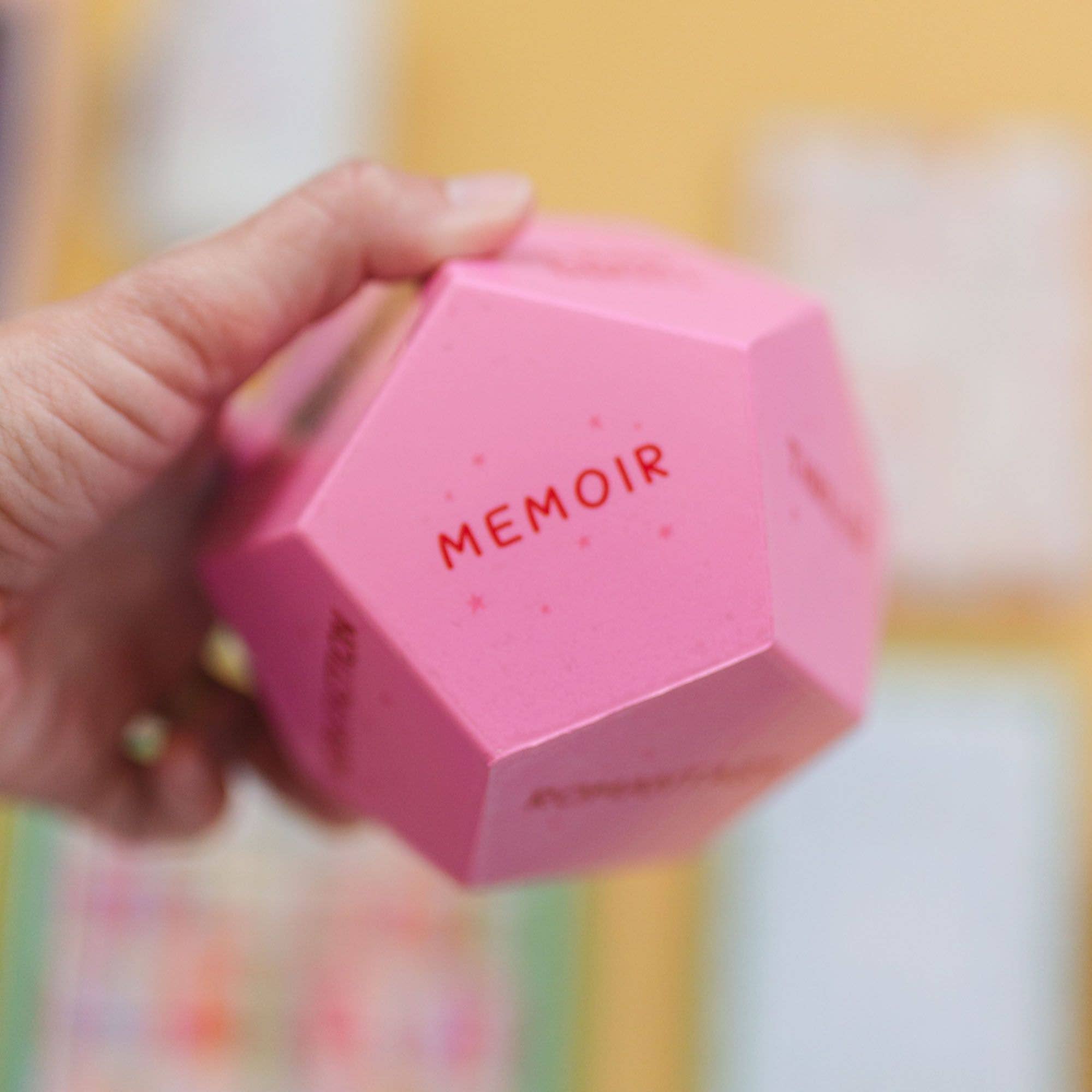 Pink hexagonal box with 'MEMOIR' text held by a hand against a colorful background