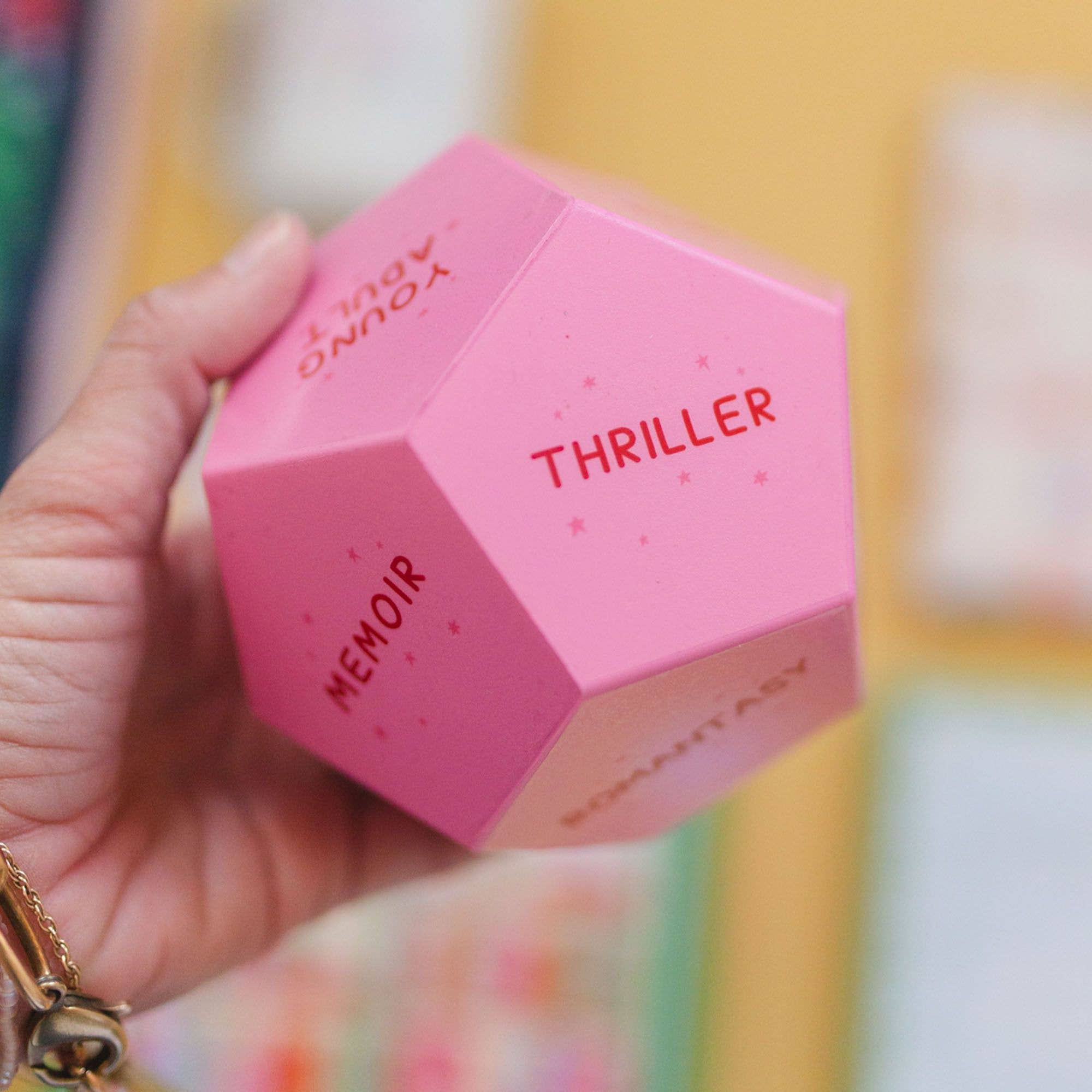 Pink hexagonal box with 'Memoir' and 'Thriller' text held by a hand against a colorful background