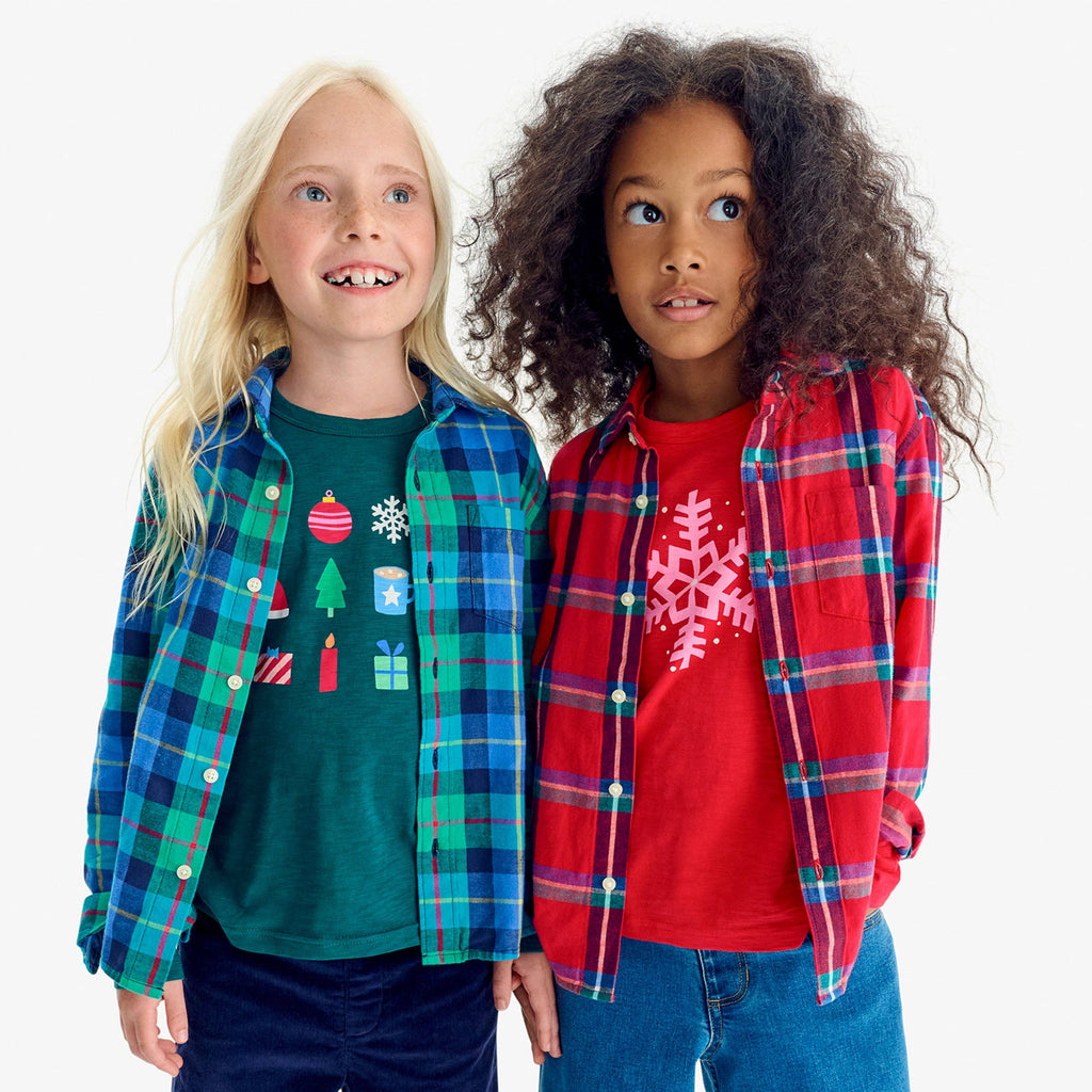 Two children modeling Christmas-themed plaid flannel shirts over graphic t-shirts, standing against a plain white studio background.