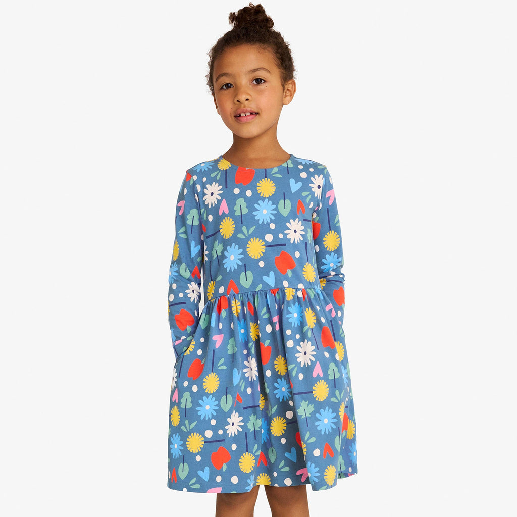 A young girl stands against a light gray studio background wearing a blue long-sleeve dress with a colorful floral and abstract print, paired with white socks and tan boots.