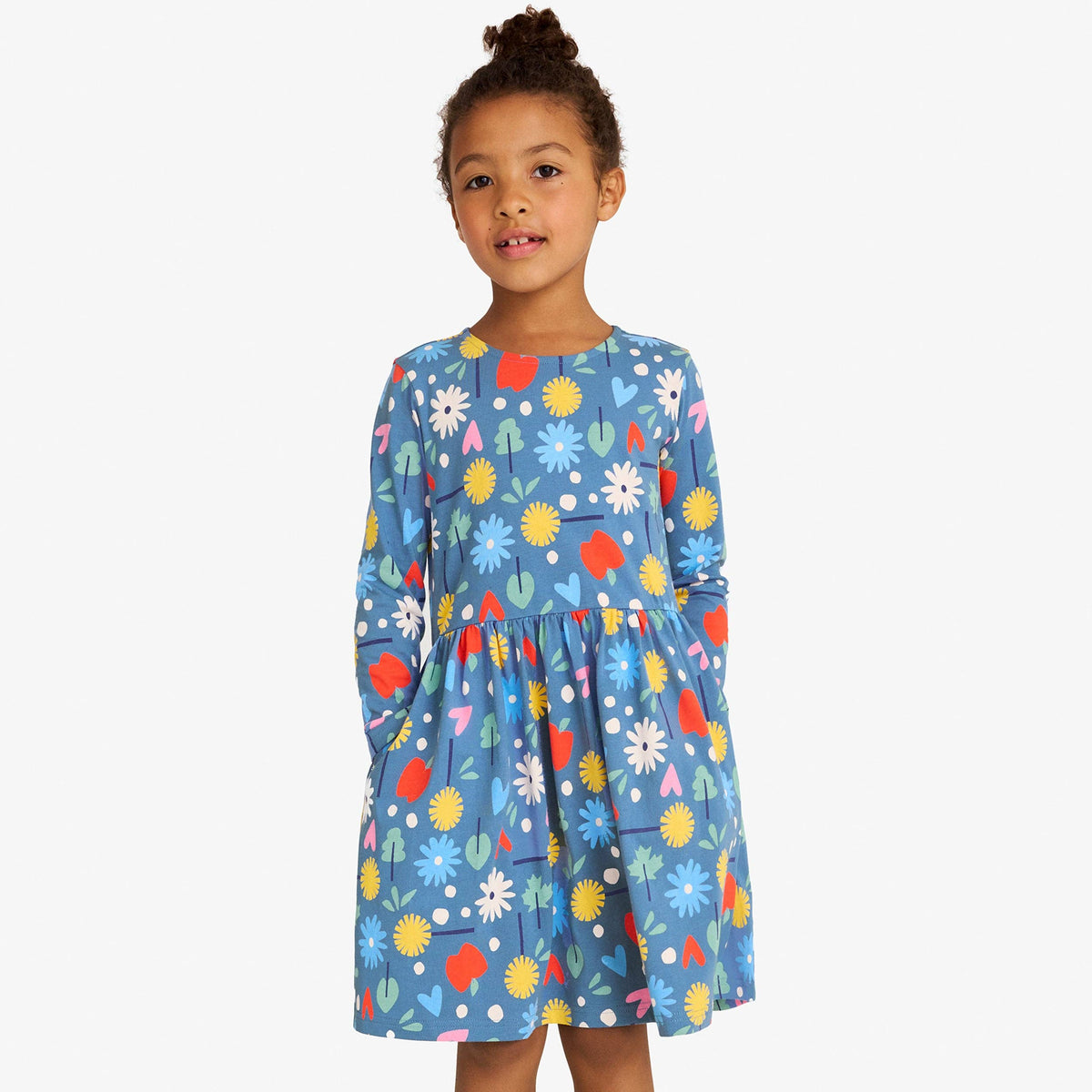 A young girl stands against a light gray studio background wearing a blue long-sleeve dress with a colorful floral and abstract print, paired with white socks and tan boots.