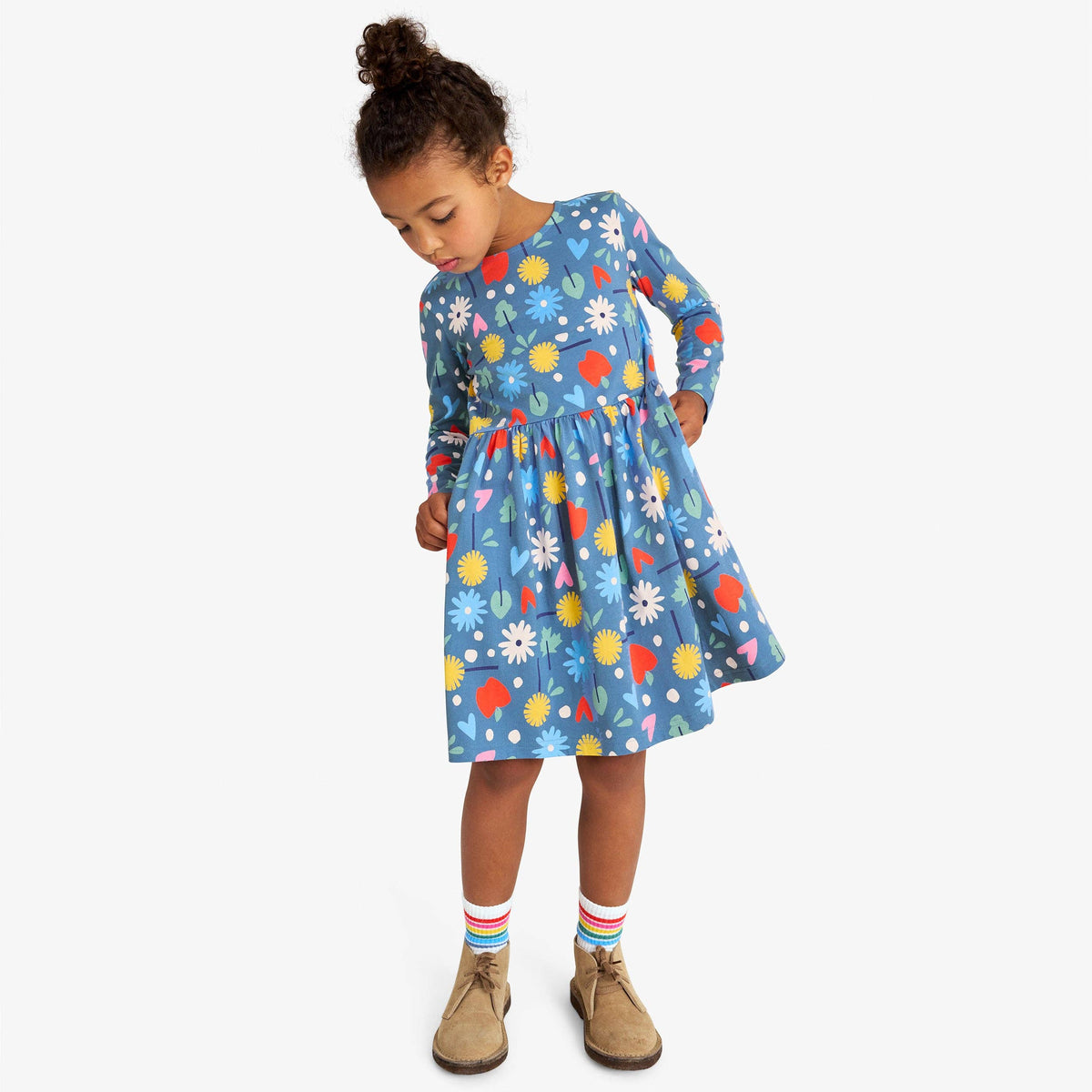 A young girl with her hair in a bun is wearing a long-sleeved blue dress with a colorful floral and abstract pattern, paired with striped socks and brown boots, standing against a light gray background.