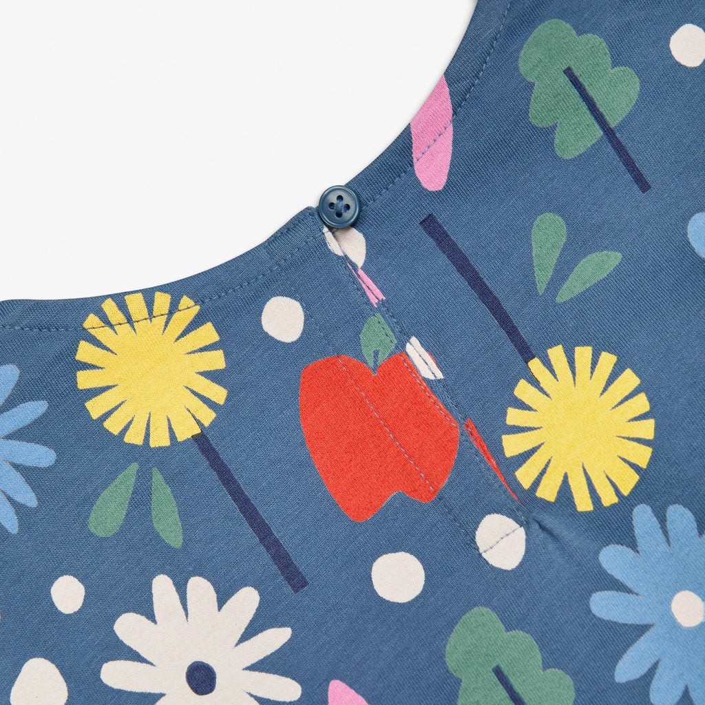 Close-up of a blue fabric garment with a colorful floral and fruit print, including yellow flowers, green leaves, a red apple, and white dots, finished with a button closure at the neckline.