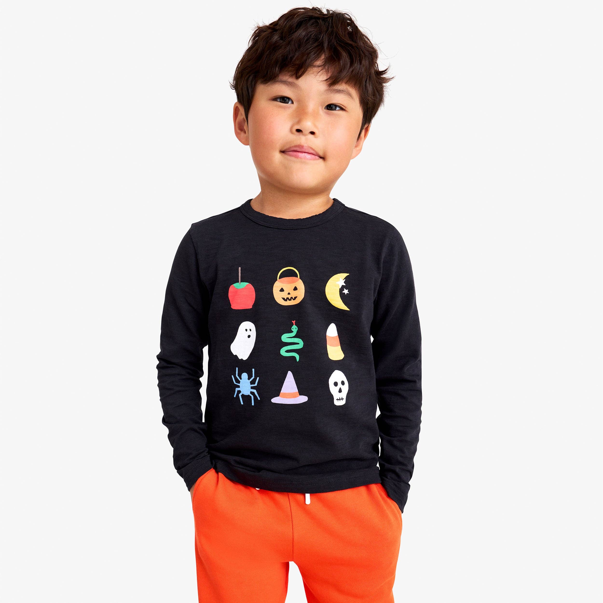 A young boy wearing a black long-sleeve crewneck sweatshirt with a colorful Halloween graphic print on the front, paired with bright orange pants, against a light gray background.