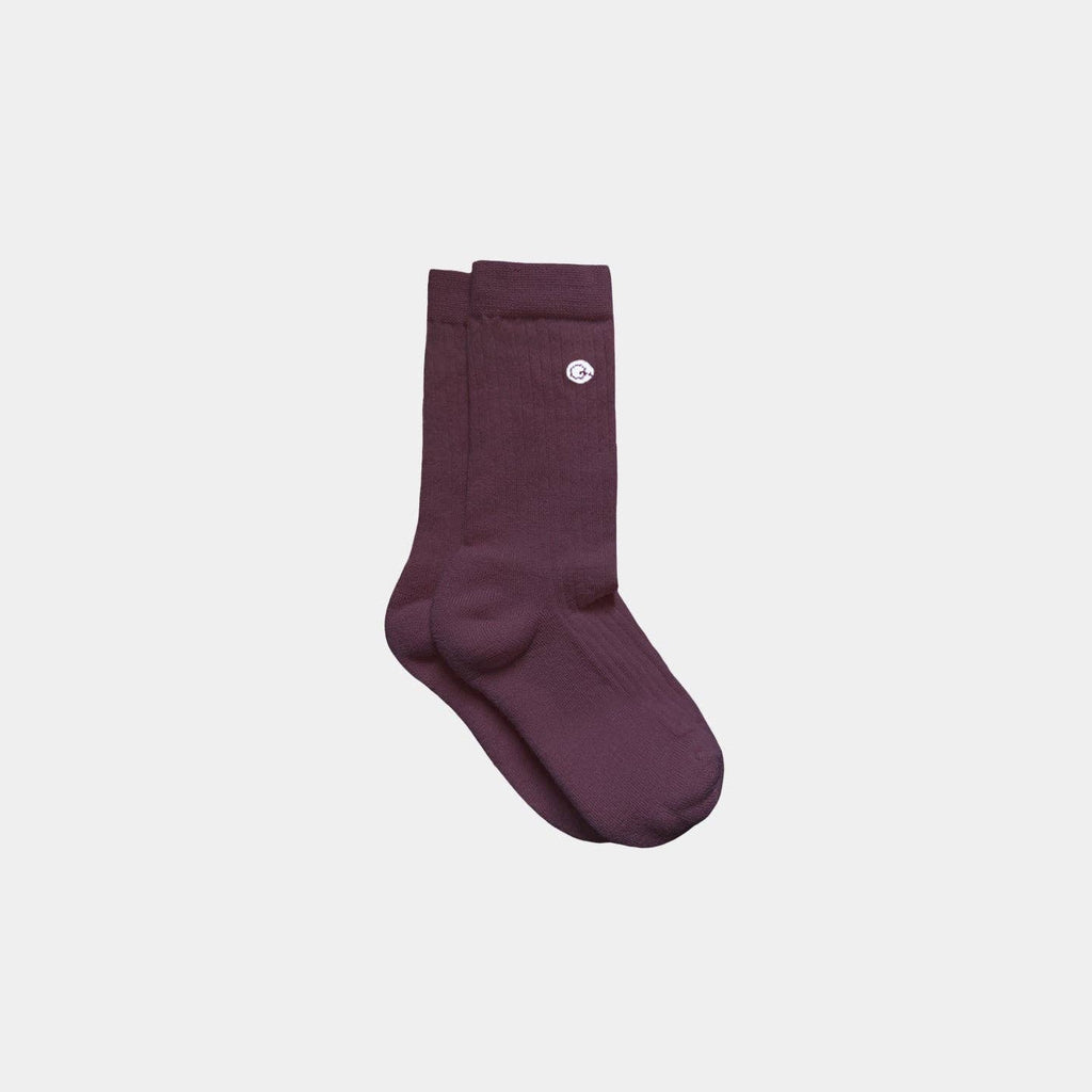 A pair of ribbed crew socks in a deep plum purple color, featuring a small white circular logo on the upper cuff, displayed against a solid light gray background.