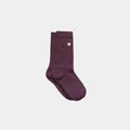A pair of ribbed crew socks in a deep plum purple color, featuring a small white circular logo on the upper cuff, displayed against a solid light gray background.