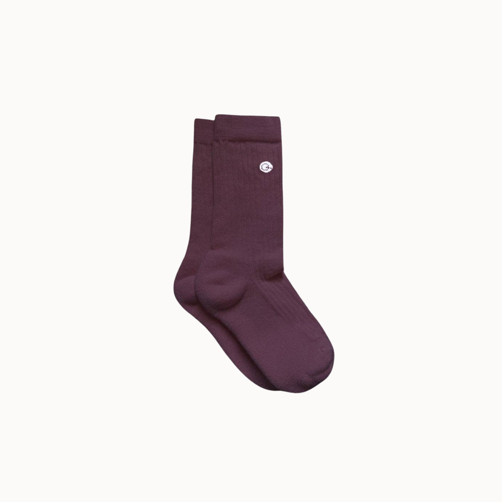 A pair of dark plum purple crew socks with a small white circular logo on the upper cuff, displayed against a solid off-white background.