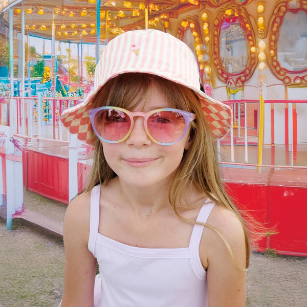 Young girl wearing a pink hat and sunglasses in front of a colorful carousel.