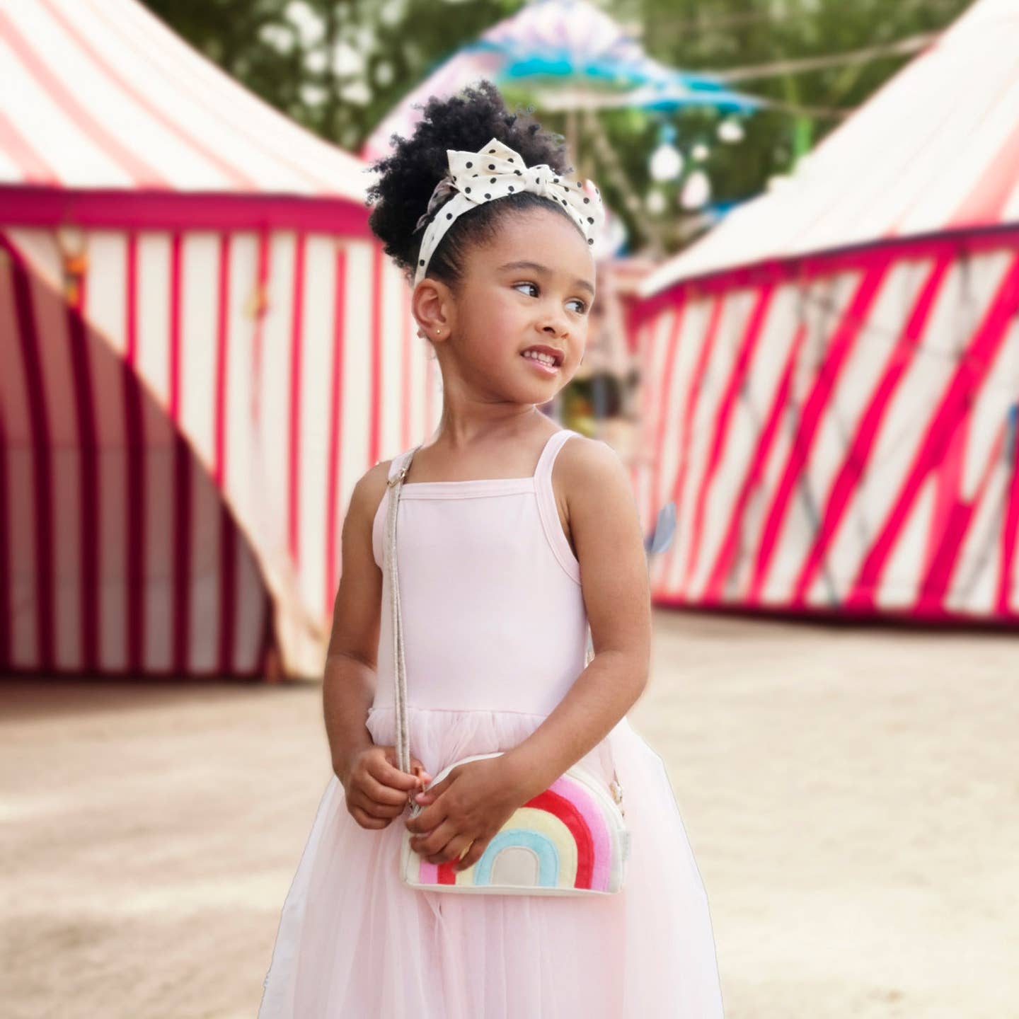 Young girl in a white dress with a rainbow bag standing in front of circus tents.