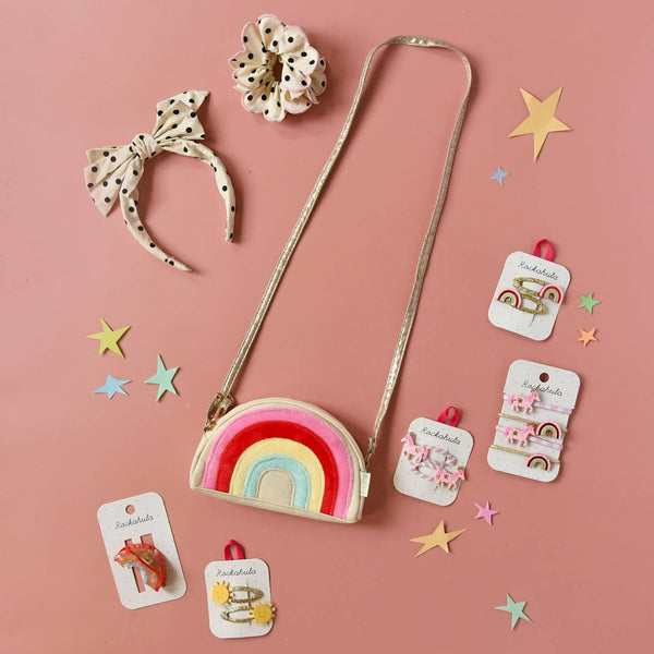 Children's accessories including a rainbow purse, headband, and hair clips on a pink background with stars.