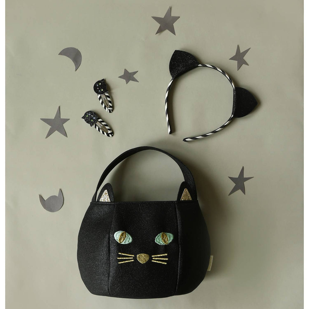 A black cat-themed fabric handbag with embroidered eyes, nose, and whiskers, displayed with matching accessories including a black-and-white striped headband with cat ears and two small hair clips on a beige background with star and moon cutouts.