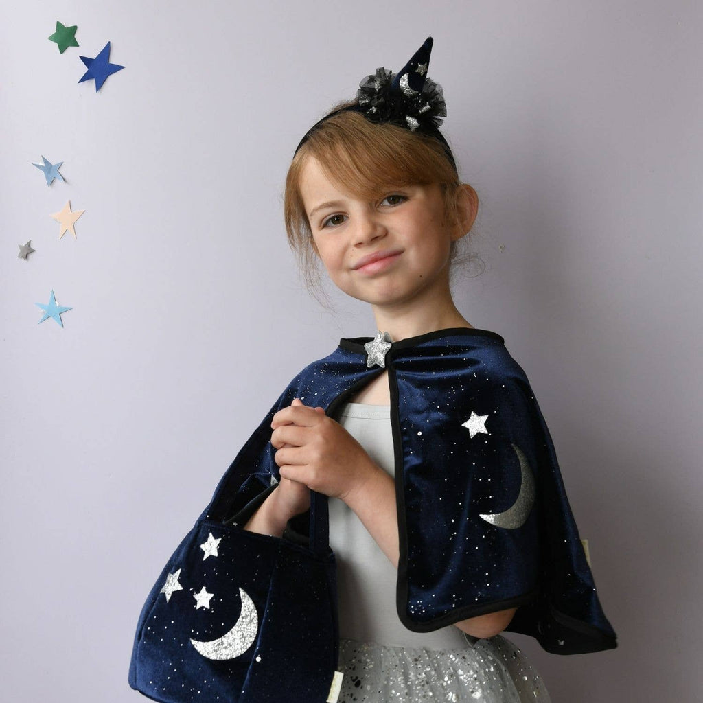 A young girl wearing a navy blue velvet cape with silver glittery stars and crescent moons, a matching black horn headband, and a sparkly silver dress, posing against a light gray wall with decorative star stickers.
