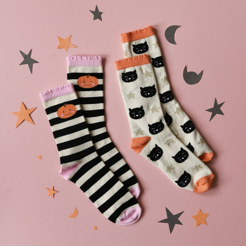 Two pairs of Halloween socks laid flat on a pink background with paper star and moon decorations. One pair has black and white stripes with an orange pumpkin and pink cuff/toe accents.