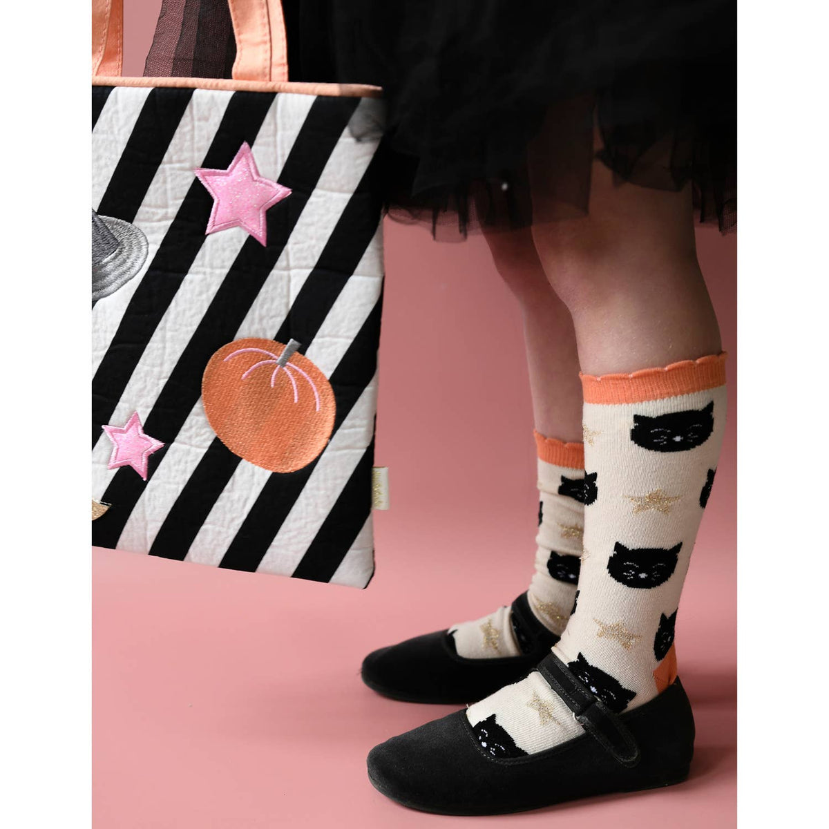 A person wearing cream socks with black cat faces and orange trim, paired with black Mary Jane shoes, holding a black and white striped tote bag with a pumpkin patch against a pink background.