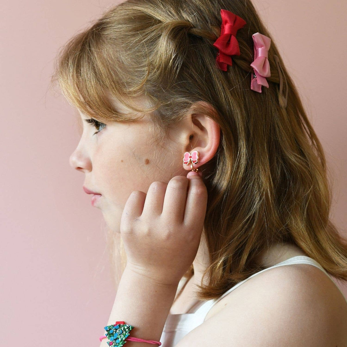 A young girl with blonde hair styled in a half-up bun with red and pink hair bows, wearing a pink flower-shaped stud earring and a colorful beaded bracelet, shown in profile against a soft pink background.
