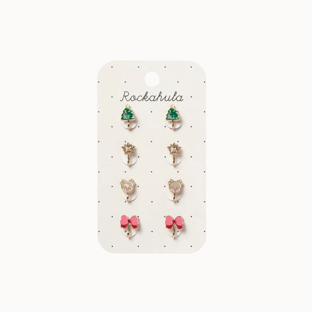 A set of six small stud earrings displayed on a white card labeled 'Rockahula'. The earrings include green Christmas trees, gold stars, pink bows, and teddy bear faces, set in gold-toned metal.