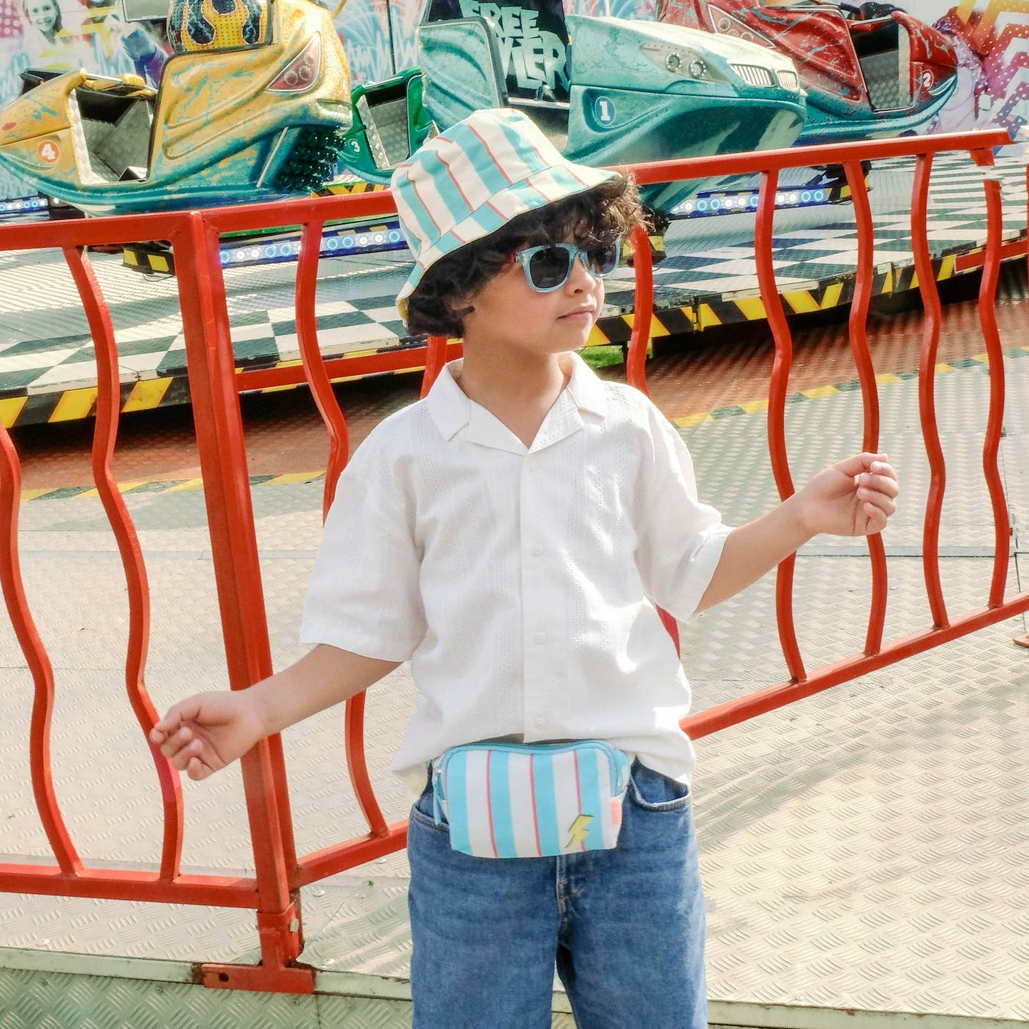Child wearing a striped hat and sunglasses at an amusement park with colorful rides in the background.