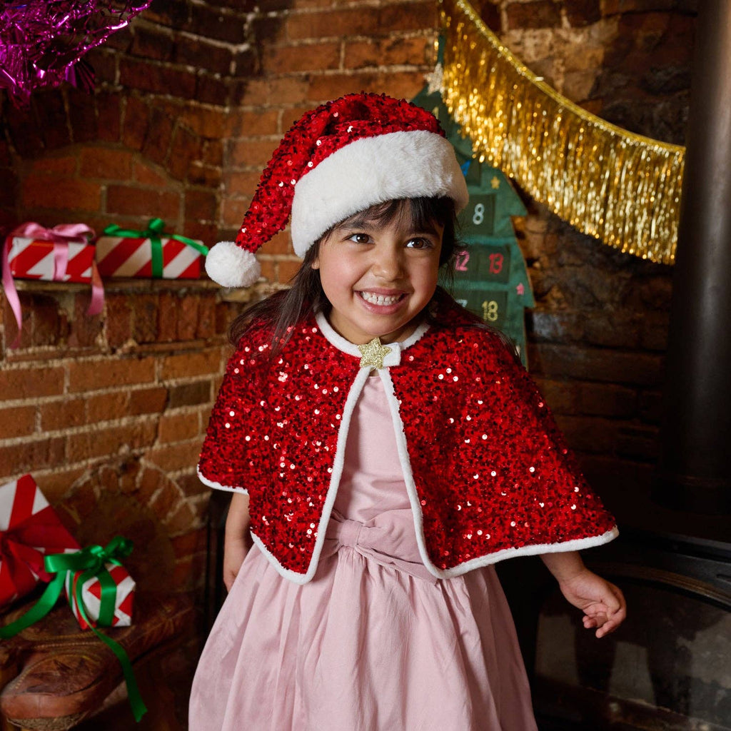 A young girl smiling in a festive holiday setting, wearing a red sequined Santa hat with white trim and a matching red sequined cape with white trim over a light pink dress. The background features a rustic brick fireplace with holiday decorations, including a gold tinsel garland and wrapped gifts.