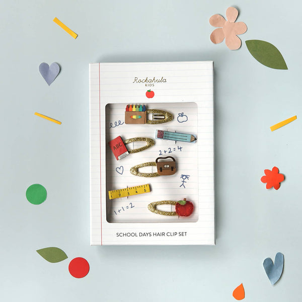 A boxed school-themed hair clip set displayed on a light blue background with scattered paper hearts, flowers, and colored sticks. The package is labeled 'Rocknroller KIDS' and 'SCHOOL DAYS HAIR CLIP SET,' showing several gold-toned clips decorated with school supplies like a pencil, ruler, notebook, and apple.