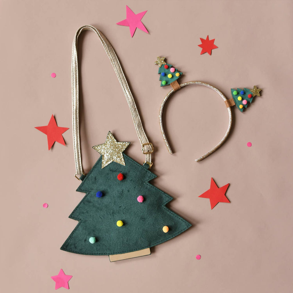 A Christmas tree-shaped shoulder bag with a metallic gold strap, decorated with a gold glitter star, colorful pom-poms, and small tree ornaments, styled on a dusty pink background with matching headband and star cutouts.