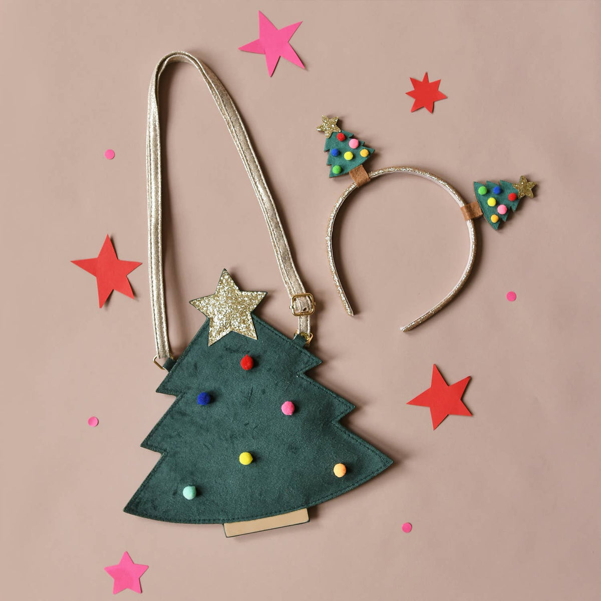 A Christmas tree-shaped shoulder bag with a metallic gold strap, decorated with a gold glitter star, colorful pom-poms, and small tree ornaments, styled on a dusty pink background with matching headband and star cutouts.