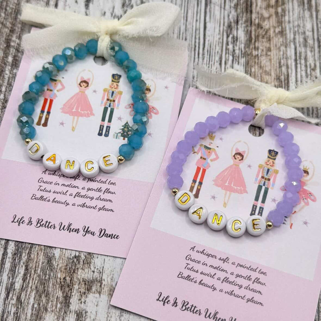 Two beaded bracelets displayed on pink cards over a rustic wood surface. One bracelet is blue and the other is lavender, both with white beads spelling 'DANCE' and white ribbon bows at the top.