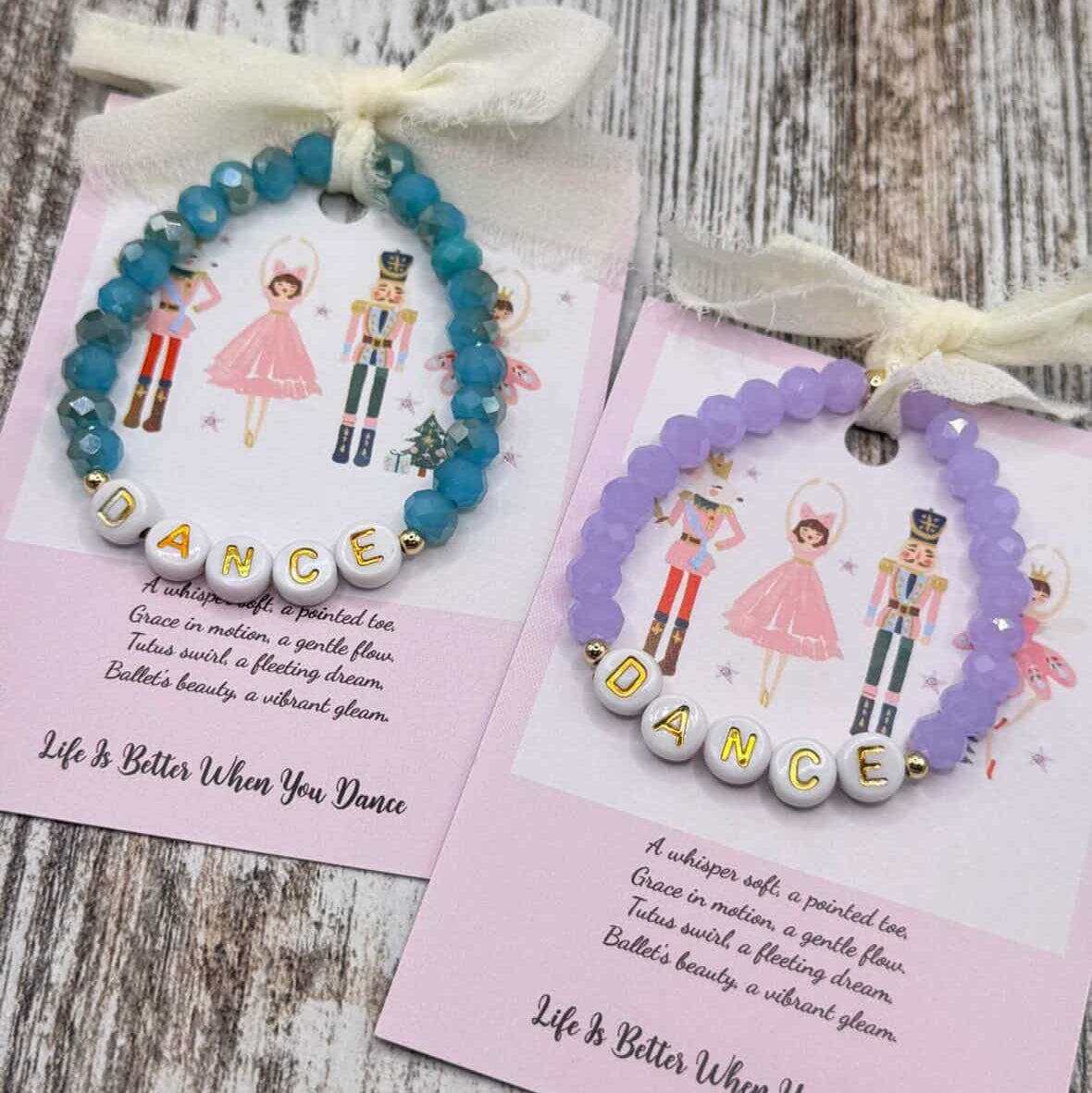 Two beaded bracelets displayed on pink cards over a rustic wood surface. One bracelet is blue and the other is lavender, both with white beads spelling 'DANCE' and white ribbon bows at the top.