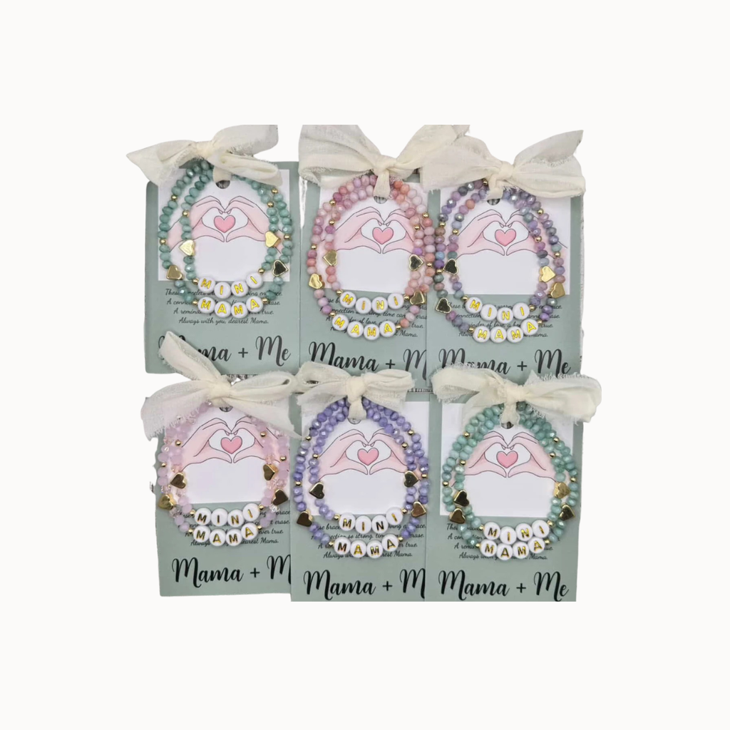 Six personalized beaded heart bracelets displayed on matching light green cards tied with white ribbons. The cards feature the text 'Mama + Me' and 'Mama + Me + 1' with heart illustrations, set against a plain white background.
