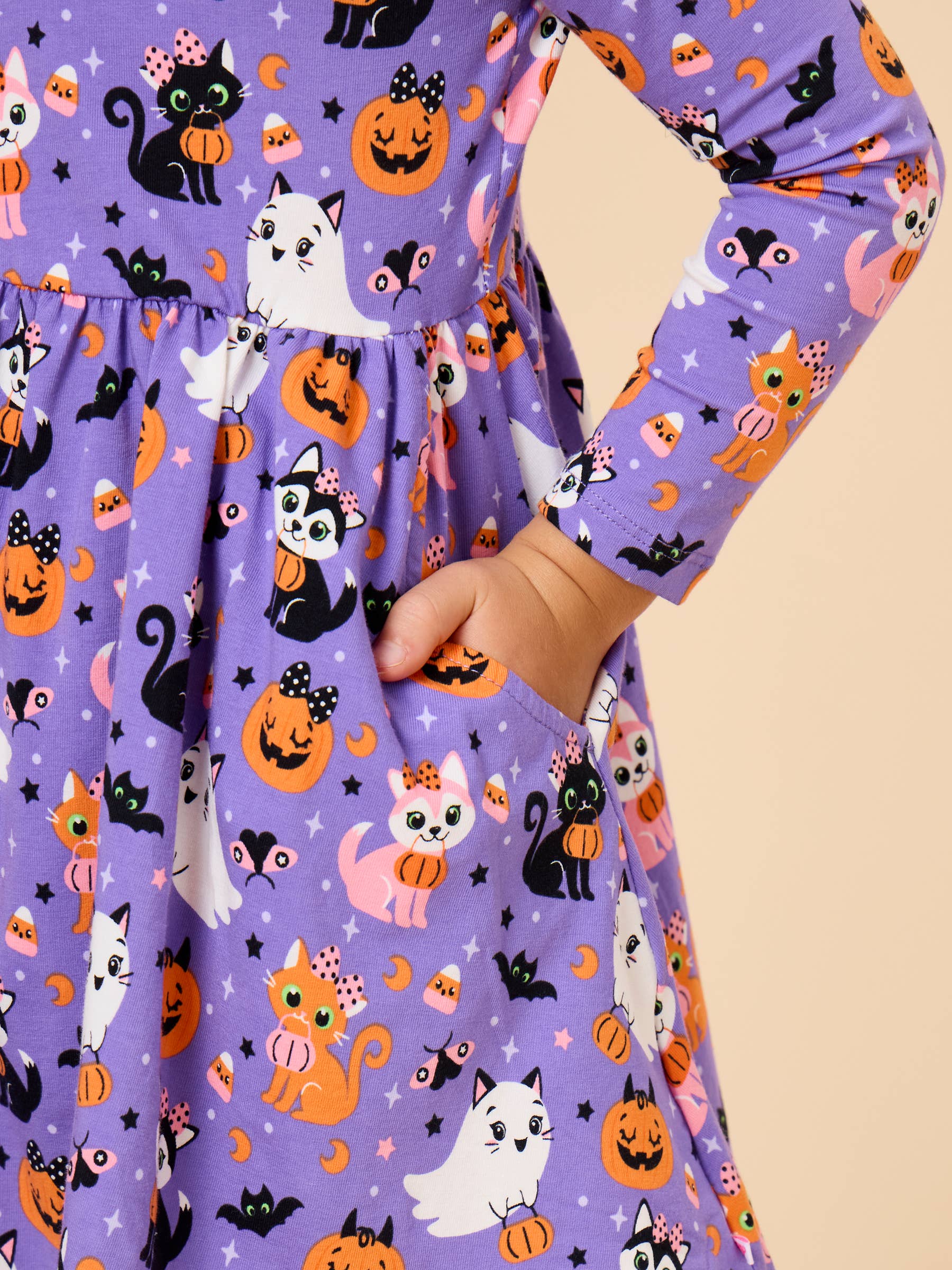 A close-up of a child wearing a purple long-sleeve dress with a Halloween print featuring black and white cats, orange pumpkins, ghosts, and bats. The child's hand is tucked into a side pocket of the dress, set against a solid beige background.