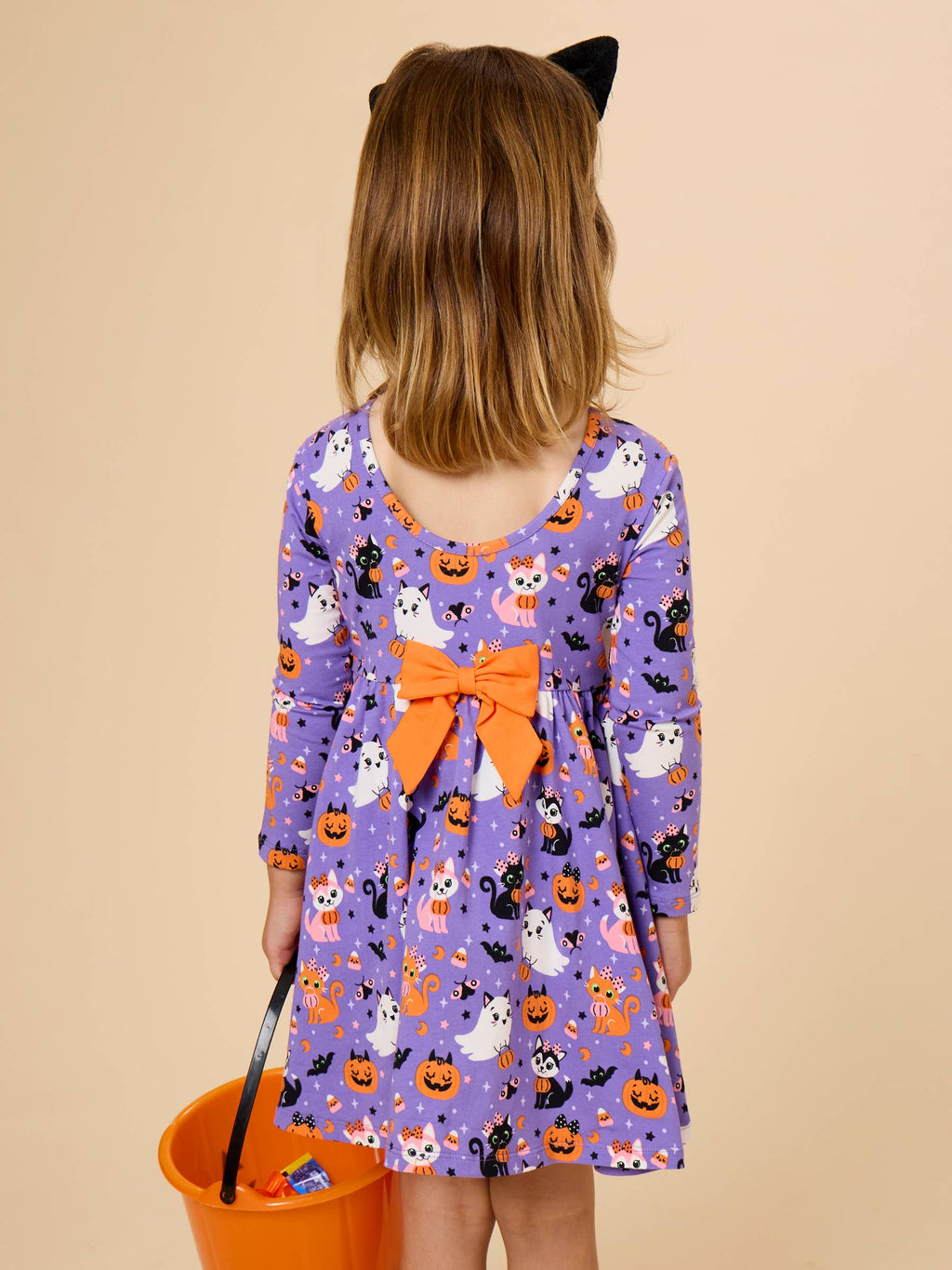 A child wearing a purple long-sleeve dress with an orange bow and a Halloween print of pumpkins, ghosts, and cats, shown from the back against a beige background.