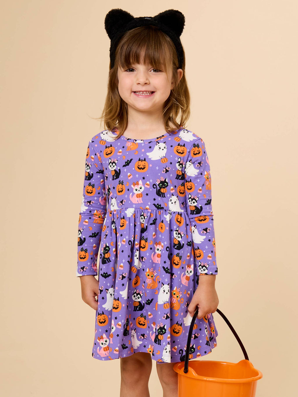 A young girl with brown hair wearing a black cat ear headband and a purple long-sleeve dress with a Halloween print of pumpkins, ghosts, and cats, holding an orange bucket against a solid beige background.