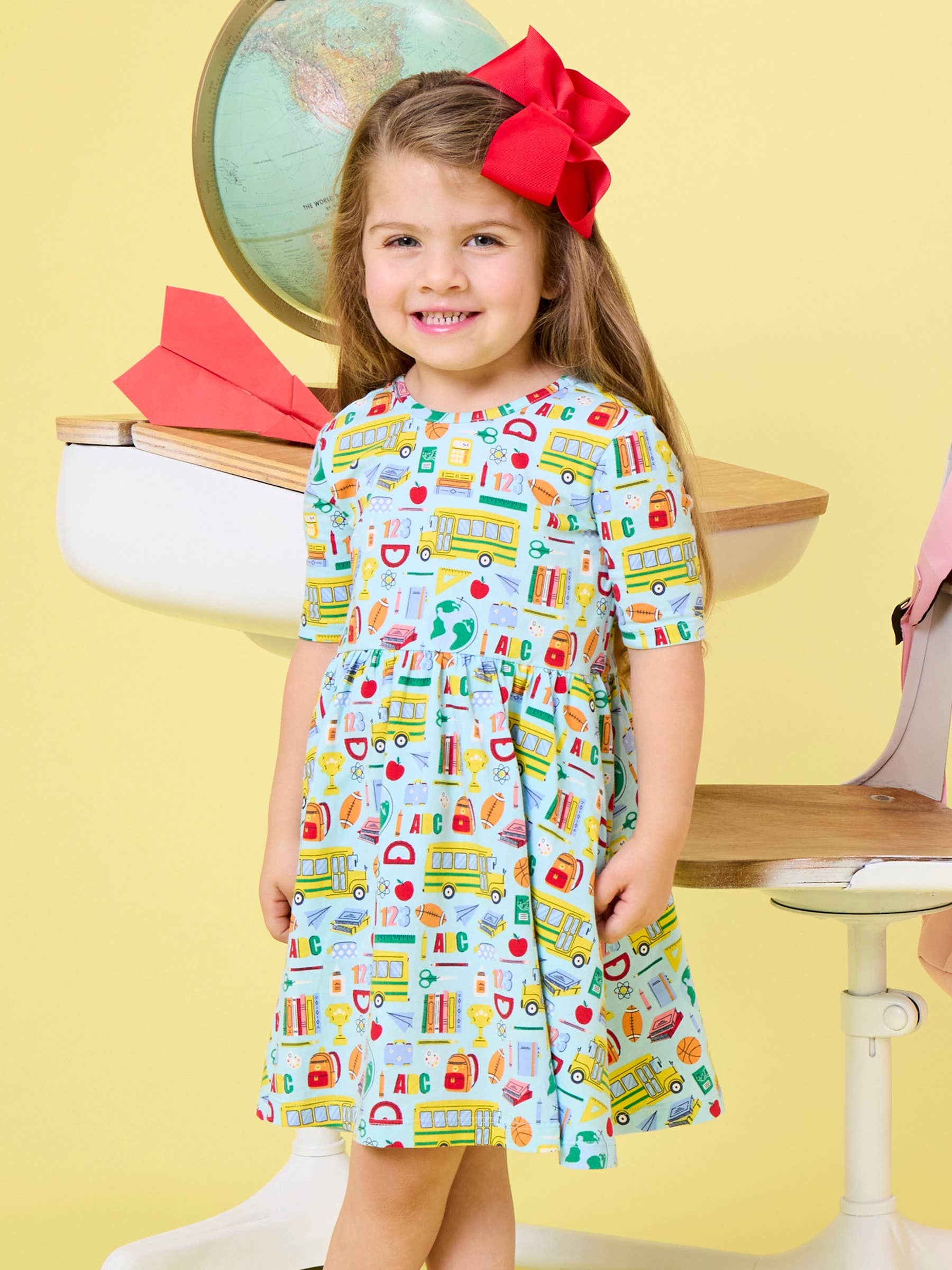 A young girl with a large red bow in her hair stands in front of a yellow background, wearing a short-sleeve dress with a colorful school-themed print of buses, books, and apples. She is positioned in front of a white desk with a globe and paper airplane prop.