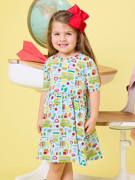 A young girl with a large red bow in her hair stands in front of a yellow background, wearing a short-sleeve dress with a colorful school-themed print of buses, books, and apples. She is positioned in front of a white desk with a globe and paper airplane prop.