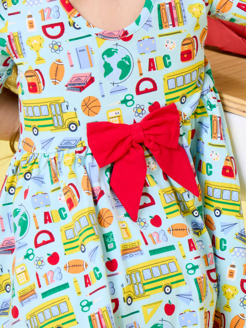 A close-up of a children's dress with a light blue background covered in colorful school-themed prints, including yellow school buses, books, globes, trophies, and red apples, with a large red bow at the waist.